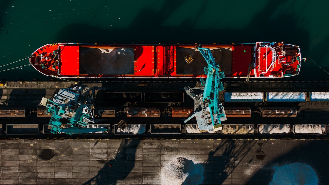 Top-down view of a bulk carrier being loaded with raw materials at an industrial dock. The ship's deck is open, revealing the vast cargo hold as heavy machinery facilitates the transfer.